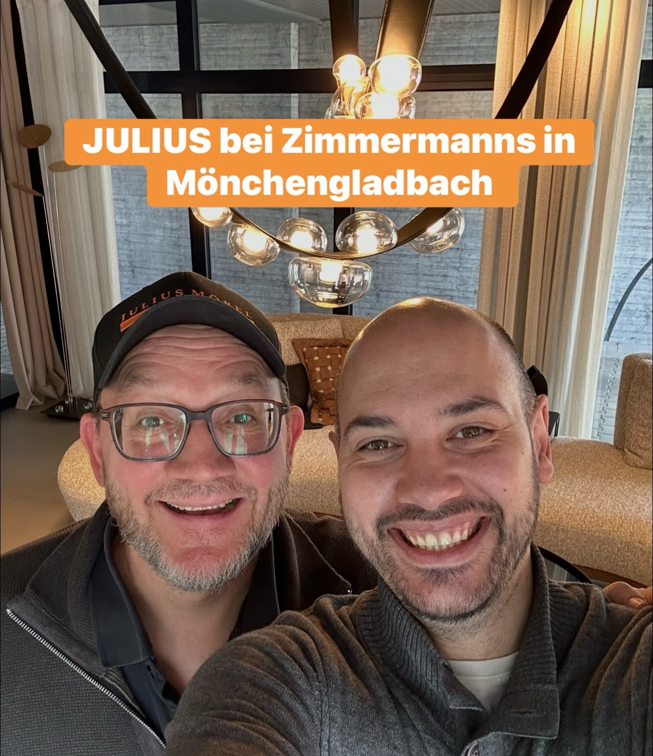 Zwei lächelnde Männer machen drinnen unter einer modernen Leuchte ein Selfie. Über ihnen steht der Schriftzug „JULIUS bei Zimmermanns in Mönchengladbach“.