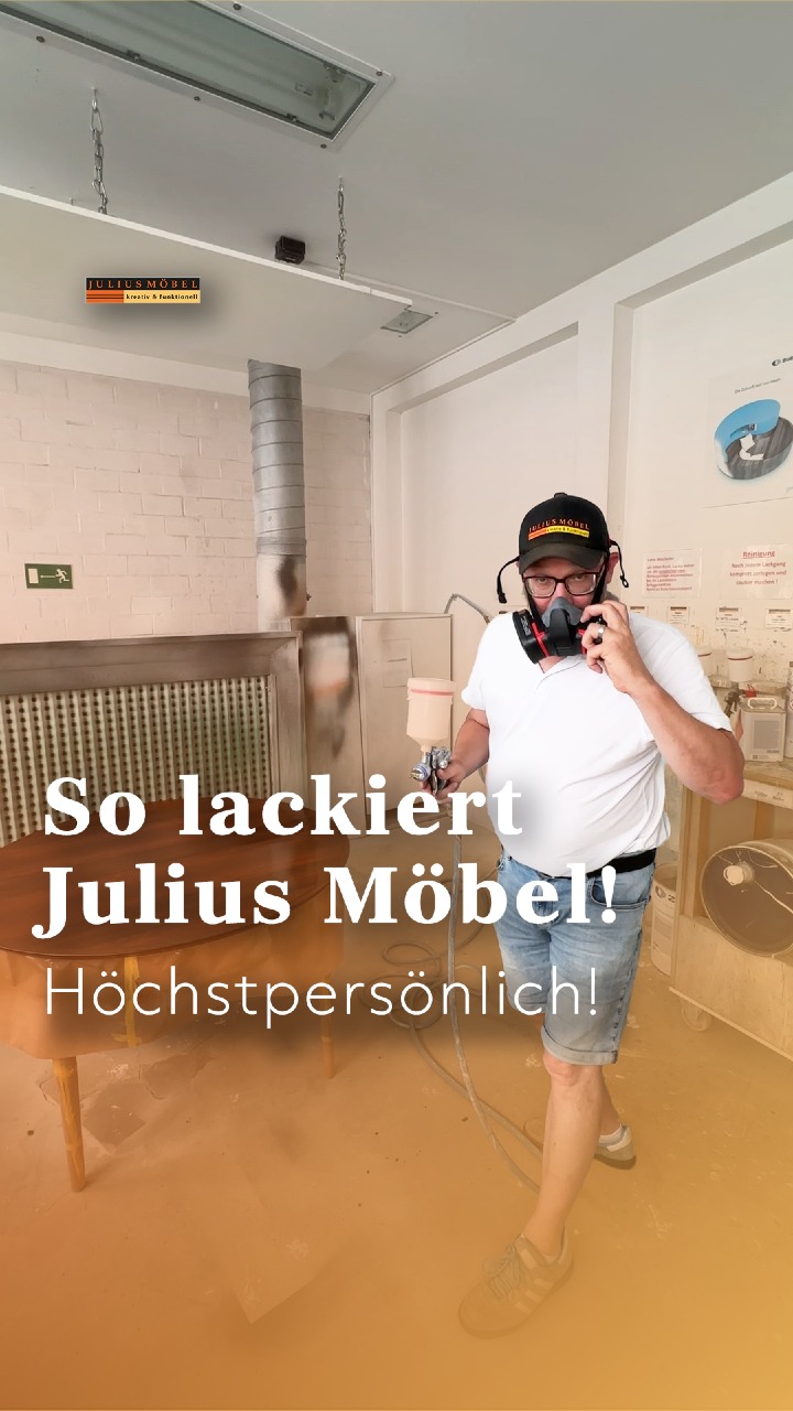 Ein Mann mit Maske sprüht in einer Werkstatt Lack auf einen Holztisch. Der deutsche Text lautet: „So lackiert Julius Möbel! Höchstpersönlich!“ Im Hintergrund sind Werkzeuge und Möbel zu sehen.