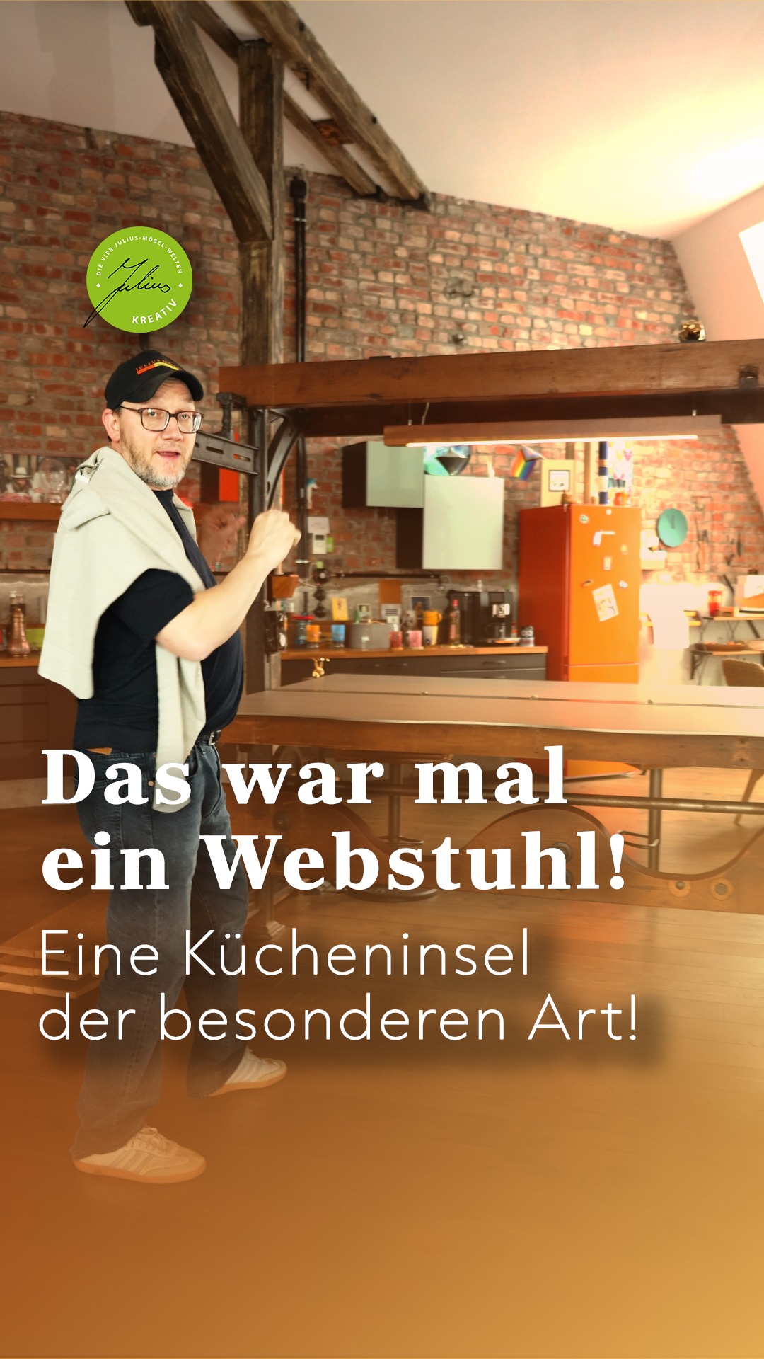 Ein lächelnder Mann gestikuliert in einer modernen Küche mit Backsteinwänden, Holzbalken und einer einzigartigen Kücheninsel. Deutscher Text: Das war mal ein Webstuhl! Eine Kücheninsel der besonderen Art!.