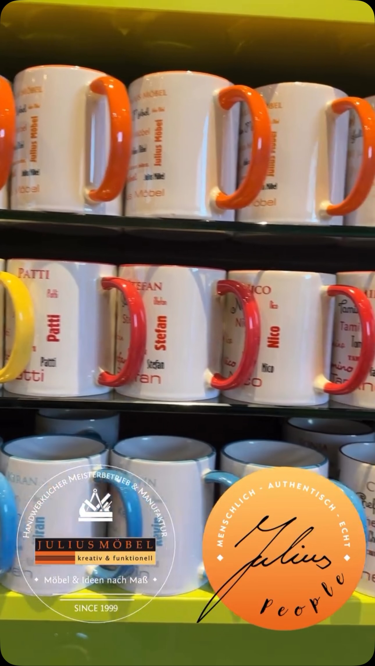 In den Regalen stehen weiße Tassen mit orangefarbenen Henkeln, die jeweils mit unterschiedlichen Namen in roter Schrift personalisiert sind. In den unteren Ecken des Bildes sind ein rundes Logo und ein rechteckiges Logo mit deutschem Text zu sehen.