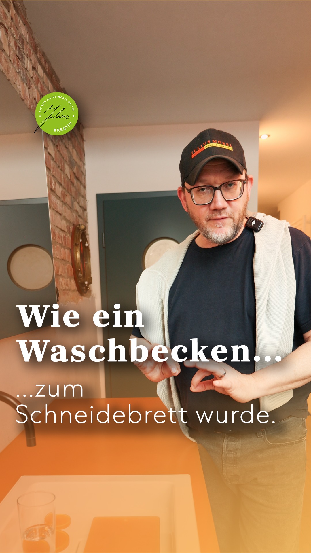 Ein Mann mit Brille und Mütze steht in einer modernen Küche und gestikuliert mit den Händen. Der deutsche Text auf dem Bild lautet: „Wie ein Waschbecken … zum Schneidebrett wurde …“.