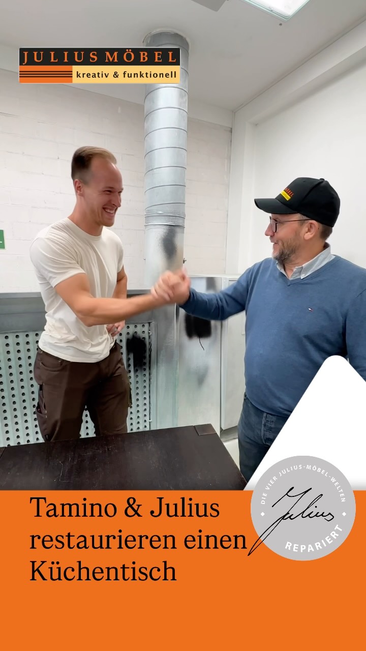 Zwei Männer, die in einer Werkstatt lächeln und sich die Hände schütteln. Auf einem orangefarbenen Banner steht „Tamino & Julius restaurieren einen Küchentisch“, in der Ecke sind das Julius-Möbel-Logo und eine Unterschrift zu sehen.