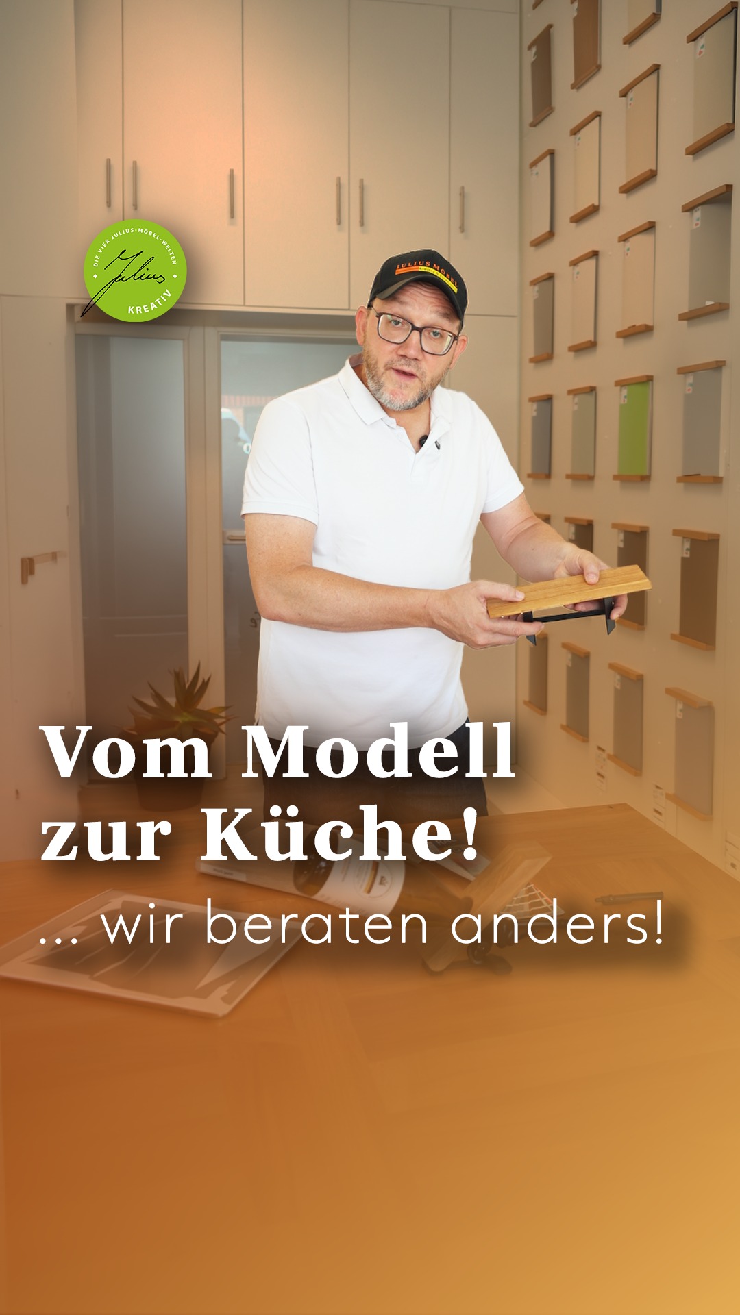 Ein Mann in einem Ausstellungsraum hält ein Muster eines Küchenschranks in der Hand. Hinter ihm sind Schränke und Muster an der Wand ausgestellt. Der Text auf Deutsch lautet: „Vom Modell zur Küche! …wir beraten anders!“.