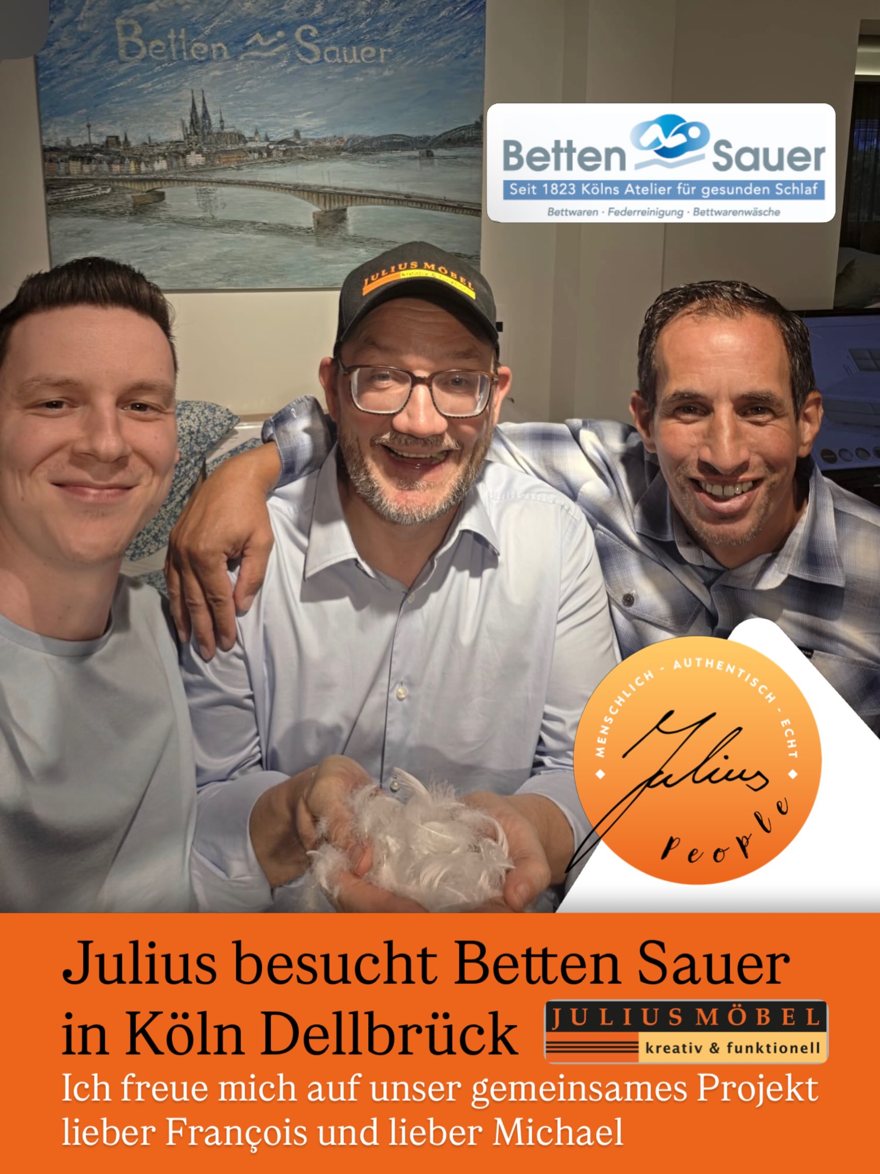 Drei lächelnde Männer posieren in einem Bettengeschäft. Auf dem Schild über ihnen steht „Betten Sauer“. Auf einem orangefarbenen Banner darunter ist zu lesen, dass Julius Betten Sauer in Köln-Dellbrück im Rahmen eines gemeinsamen Projekts mit François und Michael besucht hat.