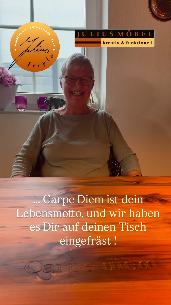 Eine lächelnde Frau sitzt an einem Holztisch, in den „Carpe Diem“ eingraviert ist. Hinter ihr sind Blumen und zwei Logos mit deutschem Text zu sehen, von denen eines die Aufschrift „JULIUS MÖBEL“ trägt. Ein Text auf dem Bild erklärt, dass das Motto für sie eingraviert wurde.