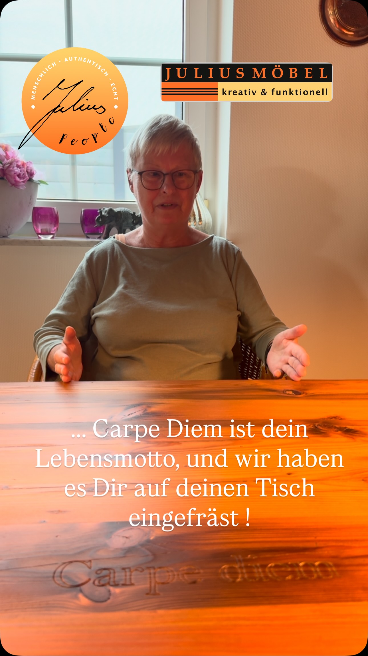 Eine ältere Frau mit Brille sitzt an einem Holztisch, in den „Carpe Diem“ eingraviert ist. Sie lächelt und streckt die Hände aus. Im Vordergrund sind Logos und deutscher Text zu sehen, im Hintergrund Blumen und ein Fenster.