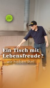Ein Mann mit Mütze und Brille steht neben einem Holztisch, lächelt und zeigt darauf. Der deutsche Text auf dem Bild lautet: Ein Tisch mit Lebensfreude? … wir haben ihn!.