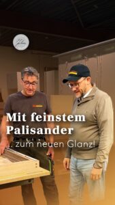 Zwei Männer in einer Werkstatt, die auf ein Möbelstück aus Holz hinabblicken; darüber steht in deutscher Sprache: Mit feinstem Palisander … zum neuen Glanz!.