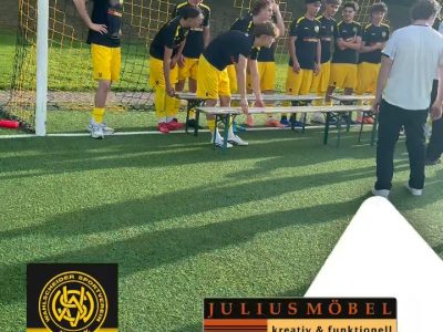 Eine Jugendfußballmannschaft in gelb-schwarzen Trikots versammelt sich auf einem sonnigen Spielfeld vor einem Tor und nimmt neue Trikots entgegen, die ein Mann an sie verteilt. Am unteren Bildrand sind Sponsorenlogos und Schriftzüge zu sehen.