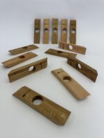 Zwölf „Balance“-Weinflaschenhalter aus Holz mit abgewinkelten Enden und einer runden Öffnung in der Mitte sind auf einer weißen Fläche zu sehen; einige stehen aufrecht im Hintergrund, andere liegen flach im Vordergrund.