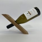 Der Weinflaschenhalter „Balance“ hält eine Weinflasche horizontal am Flaschenhals in einem Holzständer mit einem einzigen diagonalen Schlitz und sorgt so vor einem schlichten weißen Hintergrund für eine der Schwerkraft trotzende Optik.