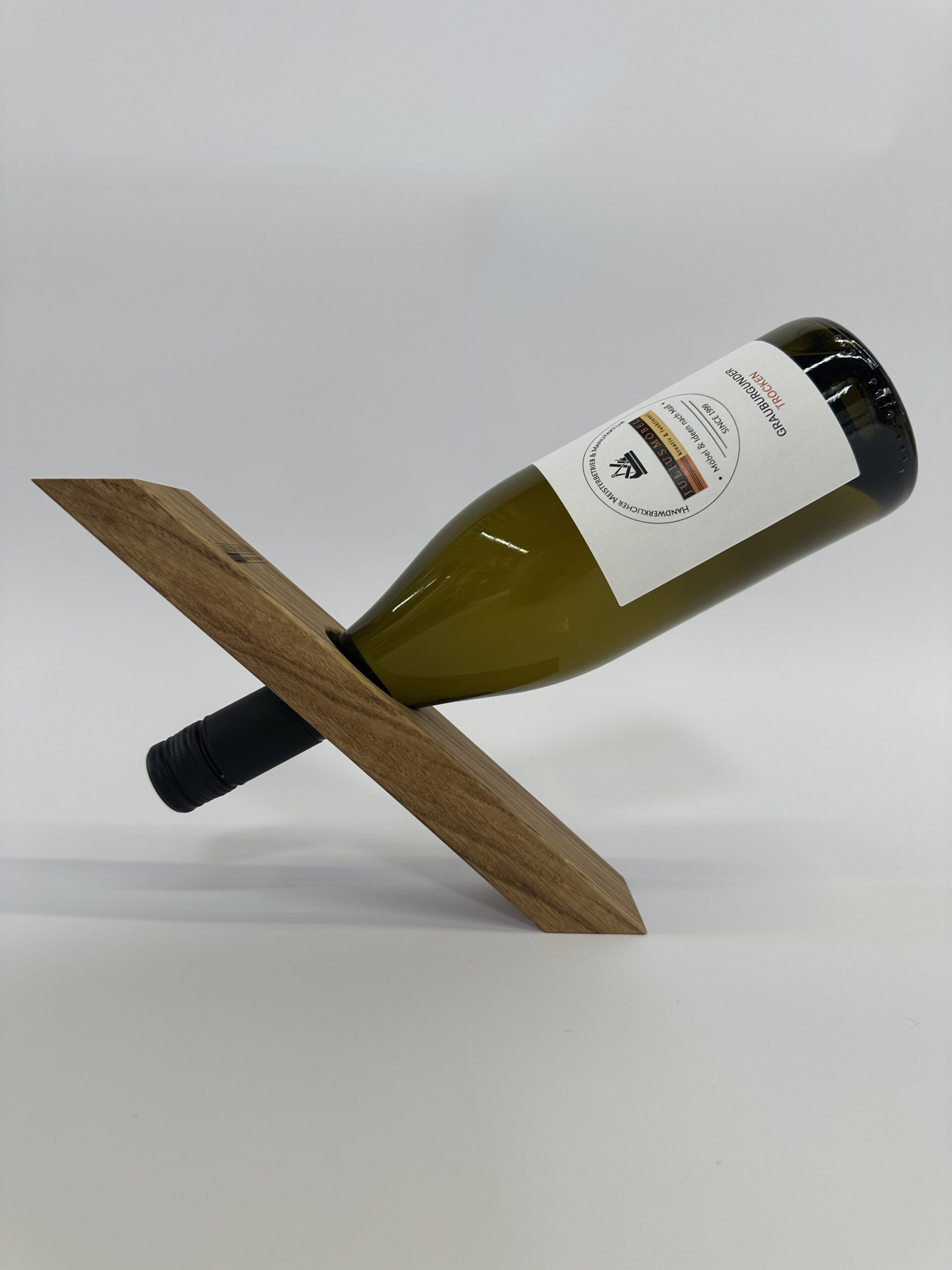 IMG_9309 Der Weinflaschenhalter „Balance“ hält eine Weinflasche horizontal am Flaschenhals in einem Holzständer mit einem einzigen diagonalen Schlitz und sorgt so vor einem schlichten weißen Hintergrund für eine der Schwerkraft trotzende Optik.
