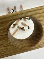 Nahaufnahme des Weinflaschenhalters „Balance“ – ein Holzbrett mit einem großen runden Loch, auf dem Miniatur-Weinflaschen und hölzerne Wäscheklammern auf einer weißen Fläche verstreut liegen.