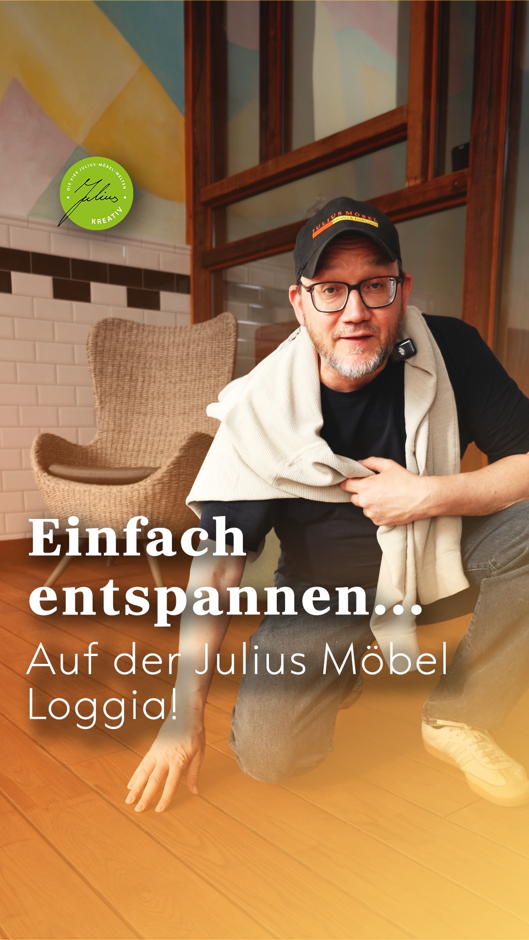 Ein Mann mit Brille und Mütze lächelt und kniet auf einem Holzboden, einen Pullover über die Schultern geworfen, neben einem geflochtenen Stuhl. Ein Text auf Deutsch wirbt für Entspannung in der Julius Möbel Loggia.