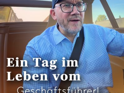Ein Mann mit Brille und Baseballkappe sitzt in einem Auto; darüber ist ein deutscher Text eingeblendet, der lautet: Ein Tag im Leben des … Geschäftsführers!.