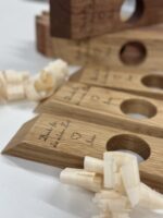 Nahaufnahme des Weinflaschenhalters „Balance“ – Holzobjekte mit runden Löchern und handgeschriebenen Botschaften, darunter ein Herz und ein Name, umgeben von Holzspänen auf weißem Untergrund. Diese personalisierten Weinflaschenhalter eignen sich hervorragend als bedeutungsvolle Geschenke oder Andenken.