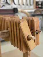 Mehrere „Weinflaschenhalter Balance“-Holzteile mit abgeschrägten Enden sind ordentlich auf einer Stange gestapelt – vermutlich als Teil eines Holzbearbeitungsprojekts – in einer Werkstattumgebung, in deren verschwommenem Hintergrund Werkzeuge und Geräte zu sehen sind.