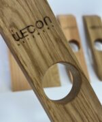 Nahaufnahme eines Weinflaschenhalters „Balance“ – ein Holzobjekt mit einem Loch und der Gravur „Uecon Netzwerk“. Im Hintergrund sind drei ähnliche Weinflaschenhalter auf einer weißen Fläche unscharf abgebildet.